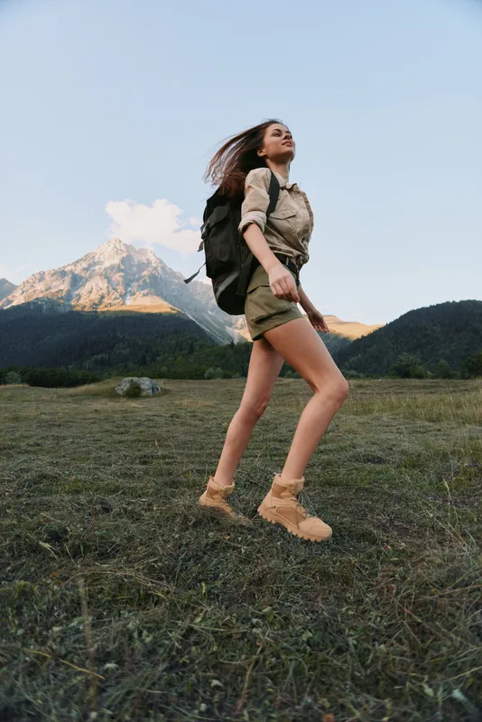 Young woman with long hair hikes on a grassy mountain trail wearing a backpack and beige hiking boots during a clear day.