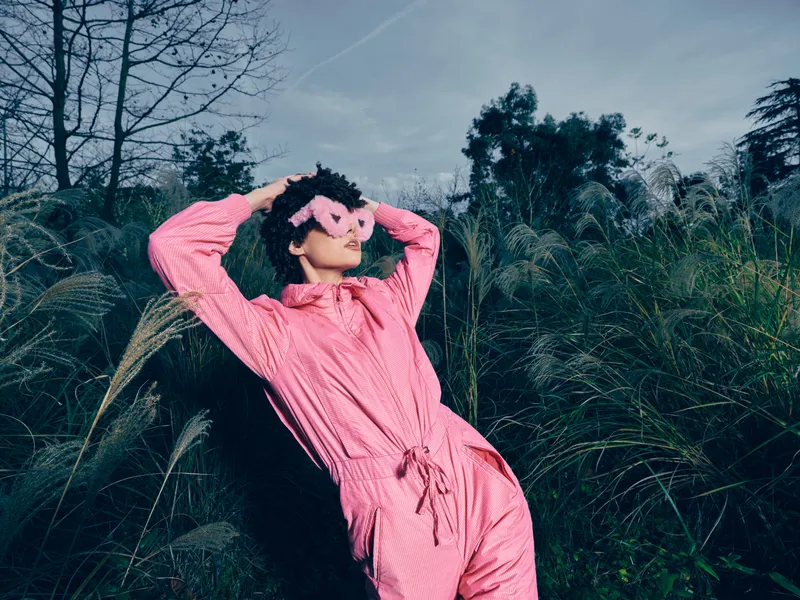 woman, pink jumpsuit, outdoor, grasses, mask, fashion, nature, playful, pose, editorial Woman lies in tall grass wearing a pink jumpsuit and playful mask posing outdoors with a dreamy vibe fashion