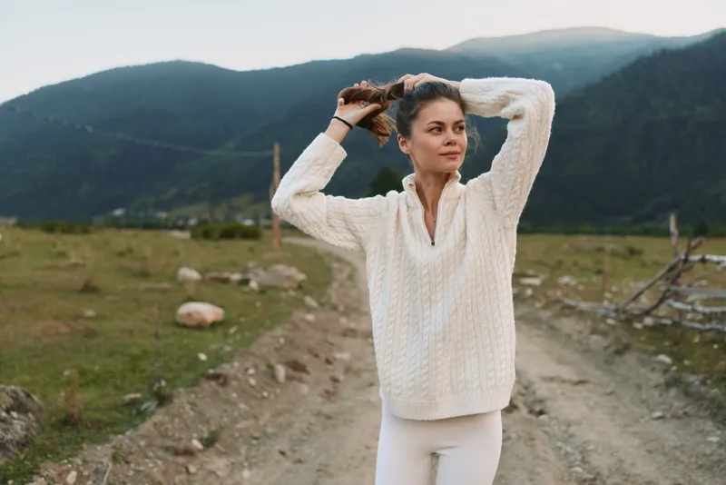 Young woman wearing a cream knit sweater stands on a dirt mountain road, adjusting her hair with rugged hills in the background, conveying a relaxed outdoor lifestyle.