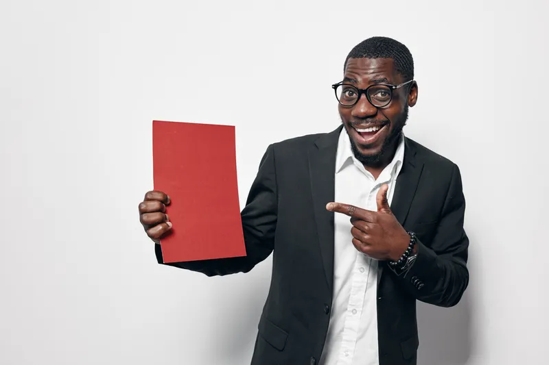 African American man with dark skin wearing glasses and black suit holding red paper and pointing at it with smile on white background. Business, presentation and confident expression.