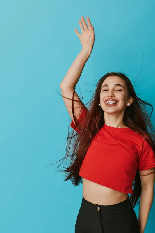 Happy woman with long hair wearing red shirt and black pants smiling and raising hand against blue background. Portrait of joyful young female with carefree expression and motion.