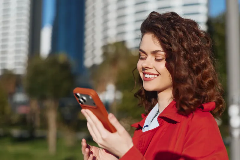 Smiling woman with curly hair checks her orange smartphone outdoors in an urban park, capturing a candid moment of everyday connectivity with bright daylight and modern buildings in the background