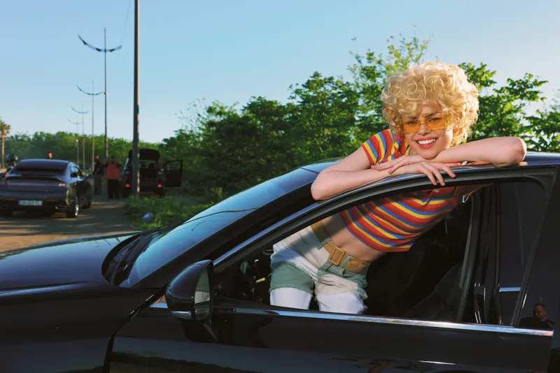 Retro woman leans from car window wearing a striped top and blond wig, bright sunny day, casual lifestyle moment with a vibrant vintage color palette