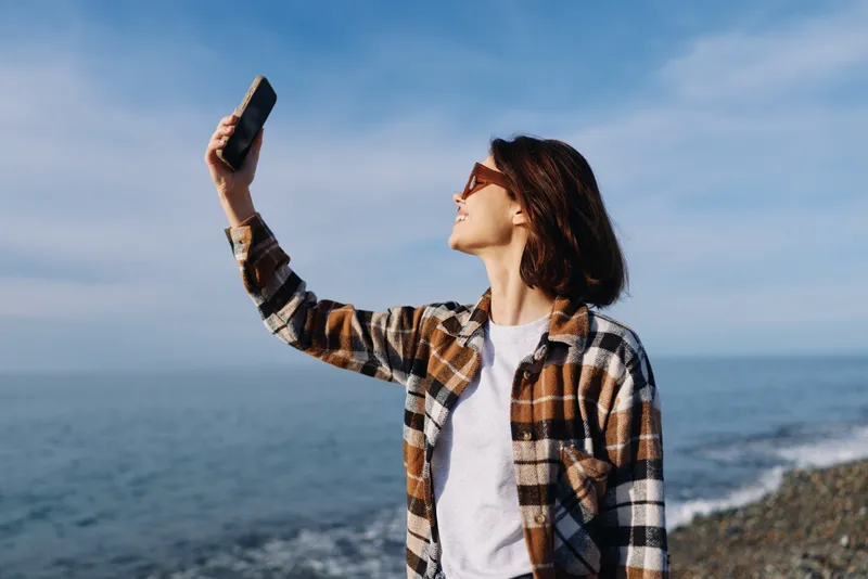 Self portrait of a woman in a plaid shirt taking a selfie with a smartphone by the seaside on a sunny day, ocean horizon, casual fashion and relaxed mood, wind in hair, coastal breeze.
