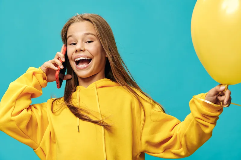 Happy girl in yellow hoodie holding balloon and talking on phone, smiling with long hair on blue background. Excited teenager enjoying conversation and celebration with bright colors.