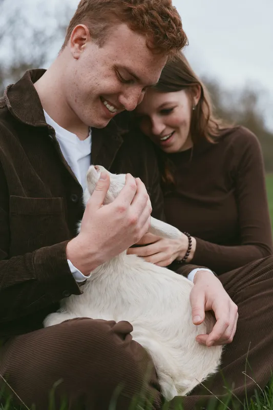A lifestyle scene shows a couple with a gentle goat kid in a grassy field, smiling and touching the animal, conveying calm, affection, and shared care in a natural rustic setting.