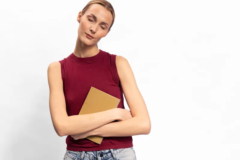 Young woman in sleeveless red top hugs closed book with closed eyes. Isolated on white background. Concept of relaxation, reading, calm, and peaceful moment.