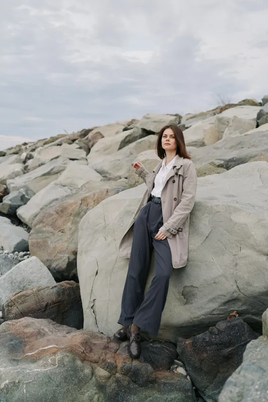 Young woman in casual outfit stands on large rocks near the coastline under cloudy sky. Concept of outdoor fashion, nature, and relaxed mood by the sea in autumn or spring season.