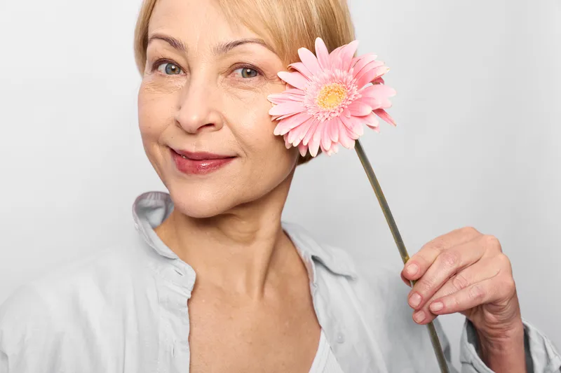 Smiling woman holding a gerbera flower near her cheek, close up portrait on white studio background, beauty wellness mood, skincare concept, self care, soft studio lighting
