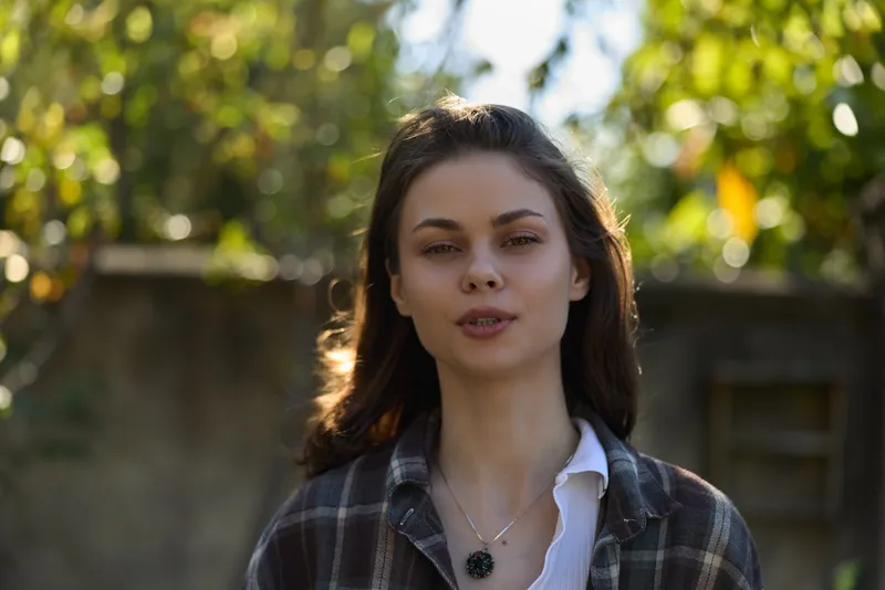 Cheerful woman outdoors, casual portrait, natural sunlight highlighting relaxed expression, plaid shirt, subtle jewelry, soft breeze, warm tones, lush garden backdrop
