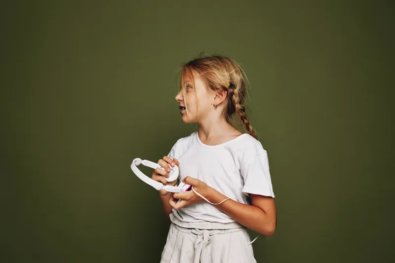 Young girl with braided hair holds white headphones and looks sideways on an olive green background. Casual outfit with a thoughtful expression. Studio portrait.