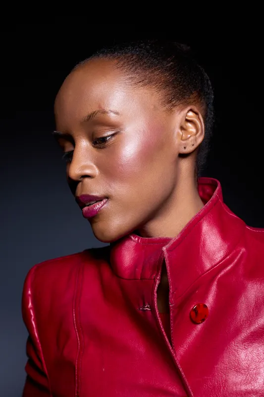 Portrait of a dark skinned African woman wearing a red leather jacket looking down with a calm expression. Studio lighting highlights her smooth skin and elegant profile.
