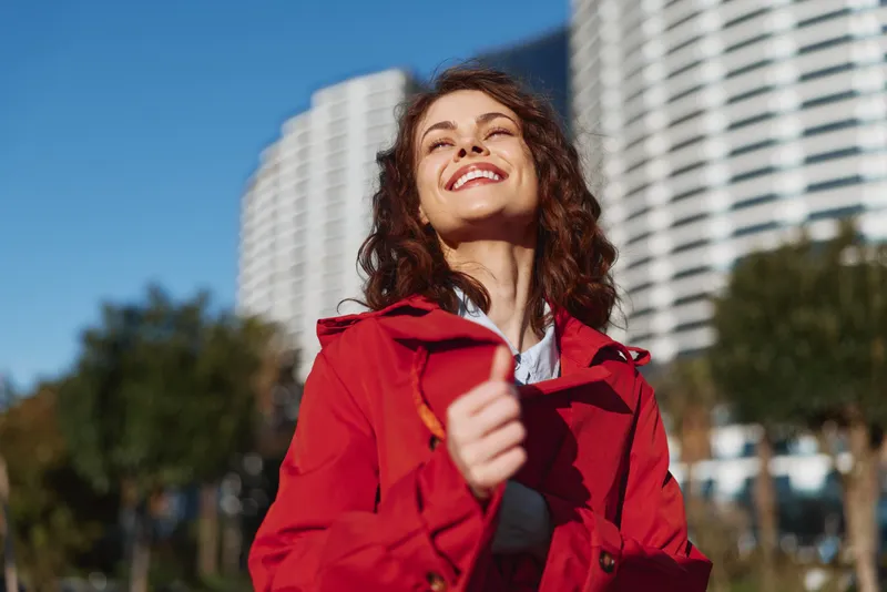 A smiling woman in a red jacket stands outdoors under bright daylight, with city buildings in the background. A vibrant moment illustrating confidence, joy, and urban energy.