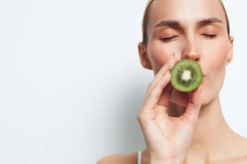 Young woman with closed eyes holding and smelling fresh kiwi fruit close to her face, studio portrait, natural skincare concept, soft lighting, white background