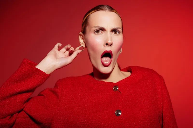 Surprised woman in red sweater pulls ear with open mouth on red background. Concept of shock, disbelief, and emotional reaction in fashion portrait photography.