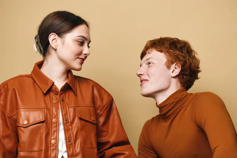 Young man and woman looking at each other with smiles, casual fashion in brown tones, studio portrait on beige background, connection and positive interaction.