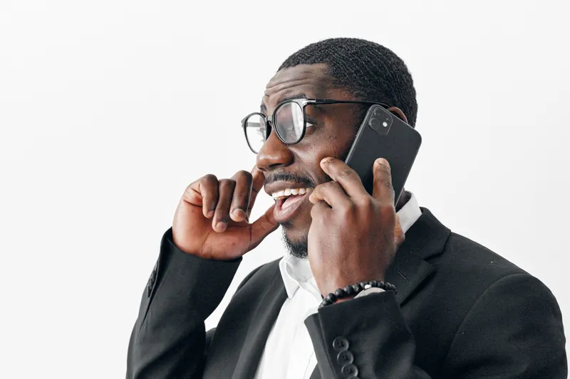 African American man with dark skin wearing glasses and black suit talking on smartphone and smiling, isolated on white background. Business communication and technology concept.