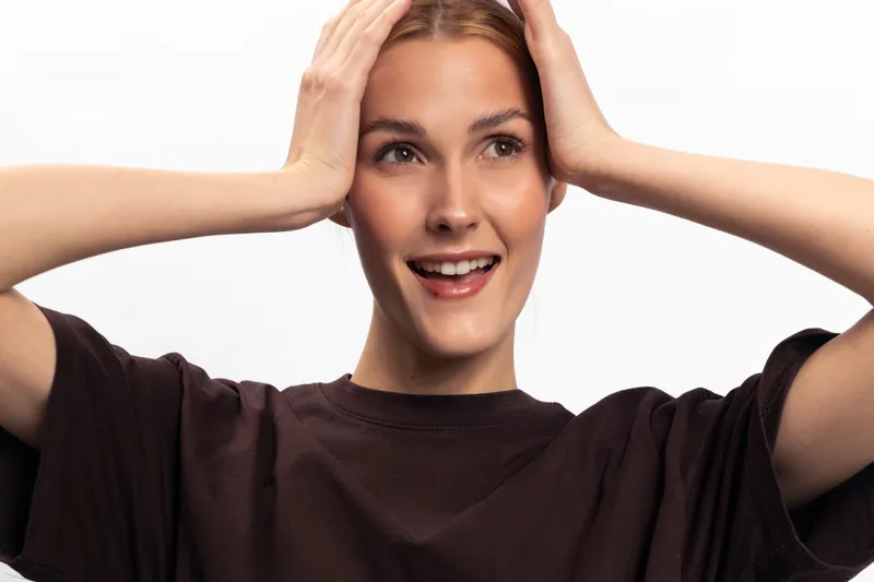 Happy young woman with natural makeup holds head with hands and smiles, isolated on white background. Concept of joy, emotions, and positive expression.