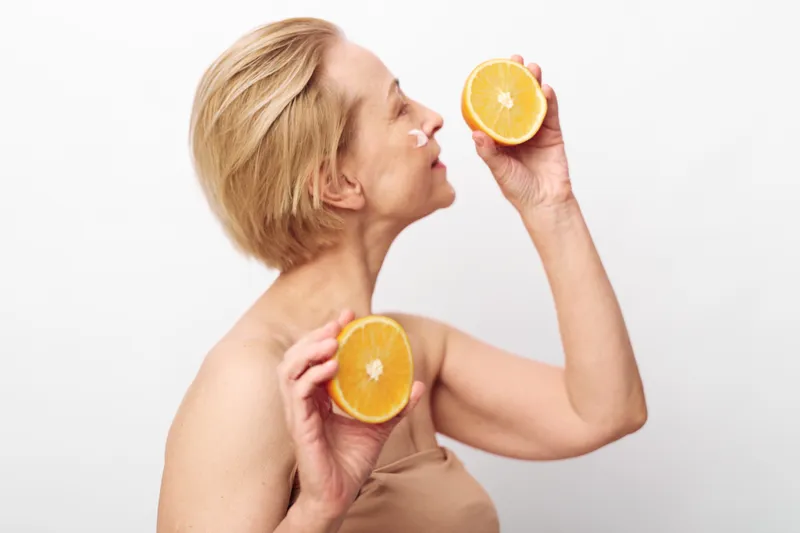 Blonde mature woman holds two halves of fresh orange near face with closed eyes. Studio shot on white background. Concept of healthy lifestyle, natural beauty, vitamin C and wellness.