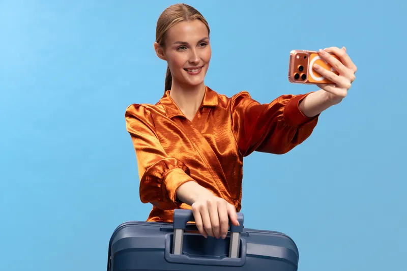 Young woman in orange blouse takes selfie while holding suitcase handle against blue background. Concept for travel, vacation, lifestyle, and social media content creation.