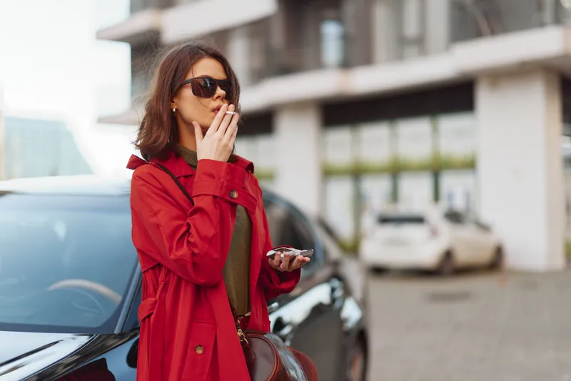 Woman wearing a red coat and sunglasses stands outdoors by a car, checking her phone, urban street scene, stylish fashion moment casual chic vibe