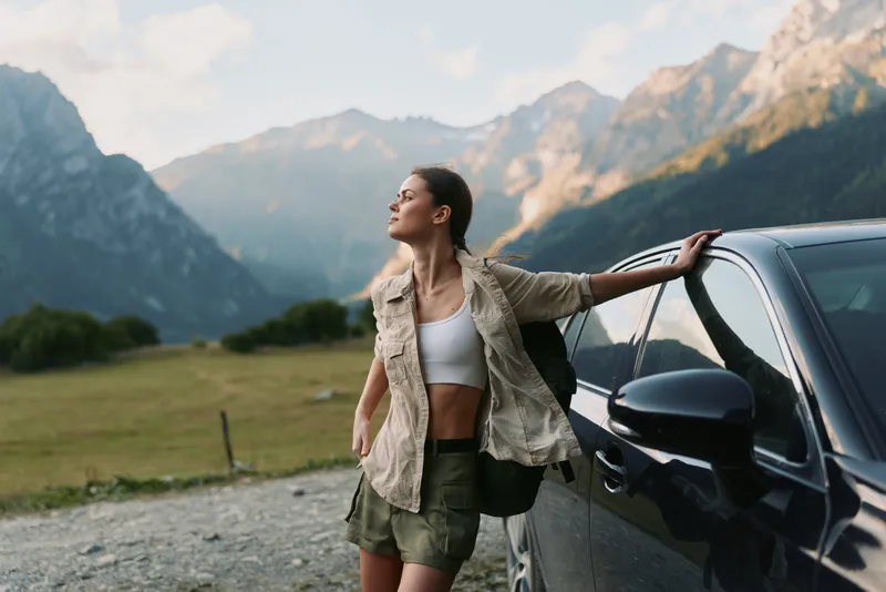 Young woman in casual outfit leaning on car outdoors with mountains in background. Concept of lifestyle, travel, freedom, and adventure in nature.