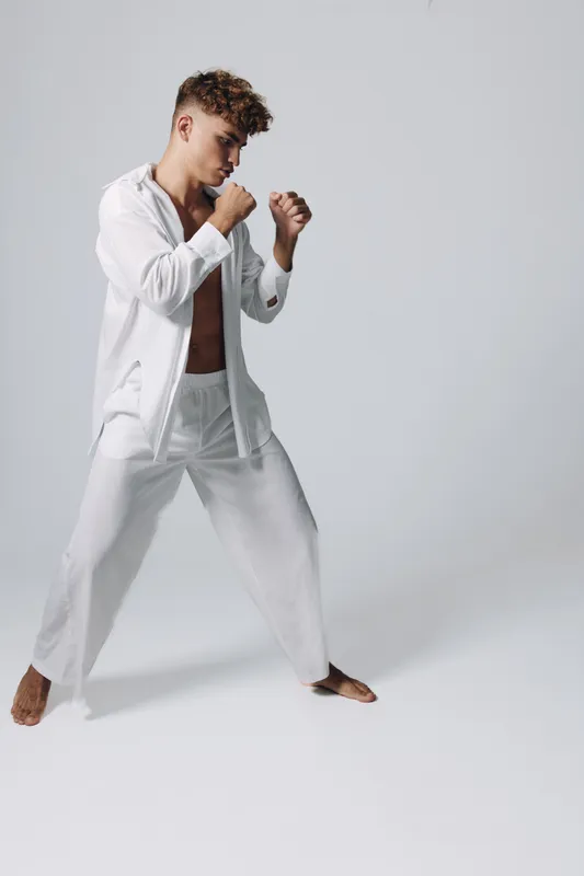 Young man in white loose clothing practicing martial arts stance barefoot on plain background. Focused athlete training with fists raised for self-defense or combat exercise.