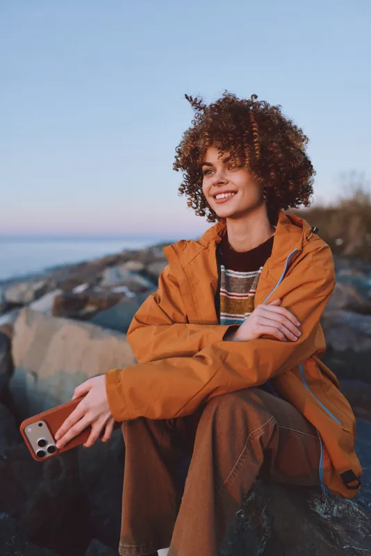 Woman lifestyle portrait outdoors, smiling, curly hair in an orange jacket, seated on rocky coast by the sea, warm sunset light, casual travel mood, relaxed pose today