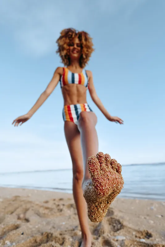 Young woman in striped bikini standing on sandy beach with foot covered in sand close to camera, bright sunny day, clear blue sky, ocean background