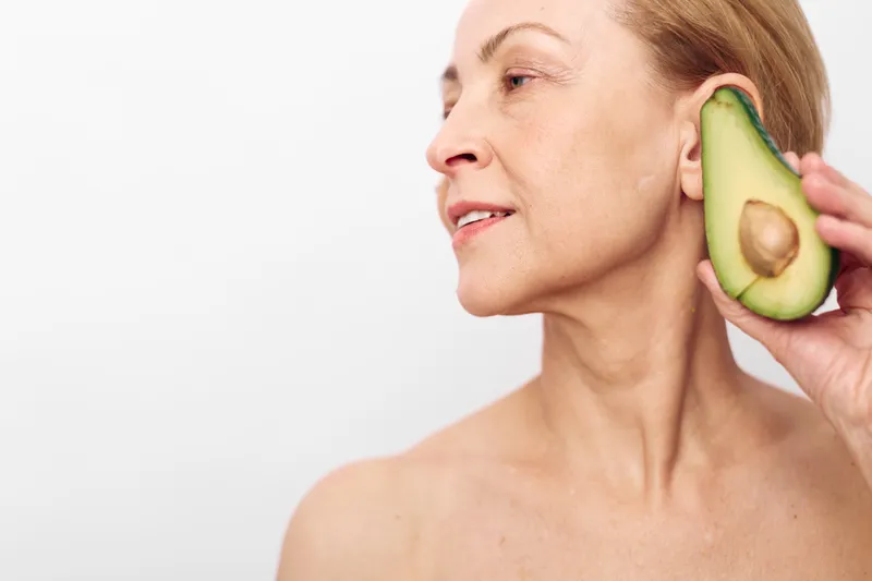 Mature woman holds half an avocado near ear, looking sideways on white background. Concept for natural beauty, skincare, healthy lifestyle, organic wellness, and self care routines.