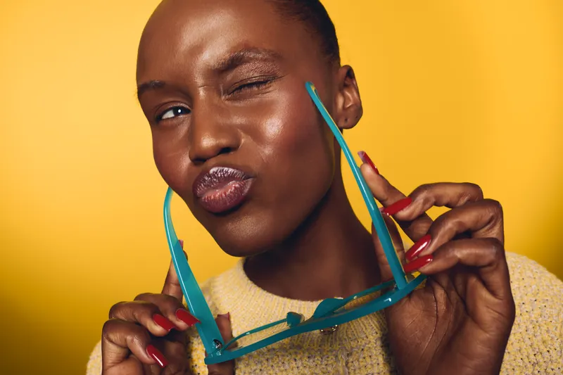 Close-up of confident dark skinned African woman with shiny skin holding blue sunglasses and winking on yellow background. Concept for beauty, fashion, and summer lifestyle.