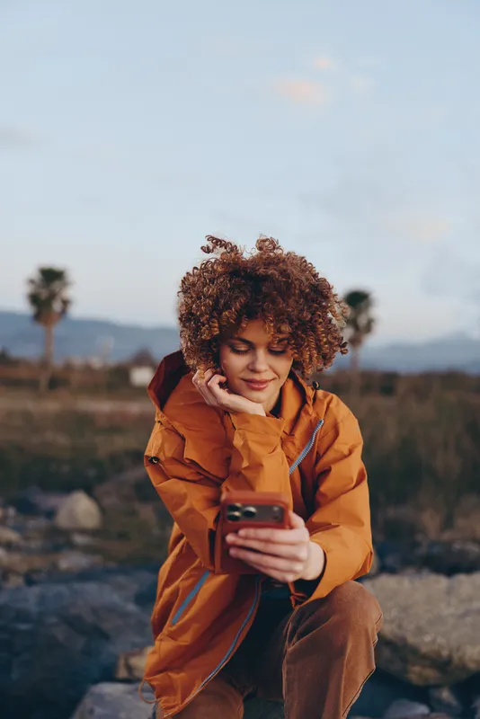 woman lifestyle curly hair smartphone outdoors jacket mountains travel smile, adult woman sits on rocky landscape, using phone in warm orange coat, relaxed portrait, natural daylight