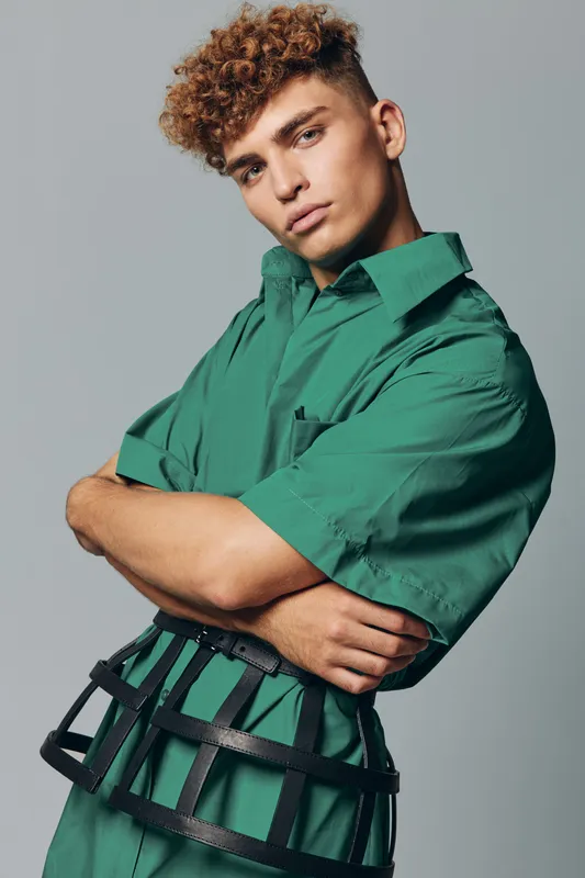 Young man with curly hair wearing green shirt and black harness posing with crossed arms against gray background. Fashion model with confident expression and casual style in studio.