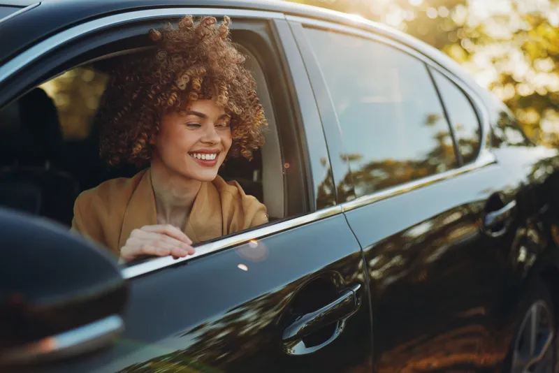 A joyful woman leans out of a parked car window, smiling at warm sunlight in a relaxed outdoor scene, capturing lifestyle moments with a retro film color aesthetic.