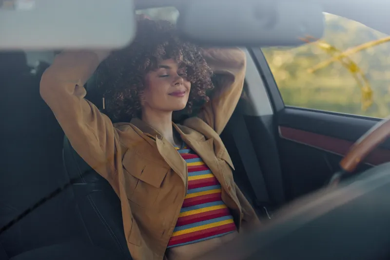 Smiling woman with curly hair sits in a car, wearing a striped shirt and retro jacket, enjoying a sunny driving. Nostalgic film color enhances lifestyle and self care.