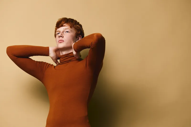 Young man with red hair wearing brown turtleneck posing with hands behind head against beige background for fashion portrait and style concept.