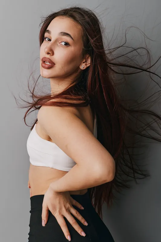 Young woman with long hair in motion posing against gray background wearing white top and black pants. Fashion model with confident expression and natural makeup in studio portrait.