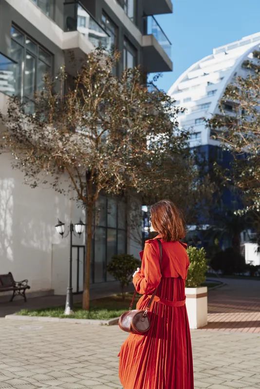 A woman in a red dress walks away in a sunny urban plaza, carrying a small round bag, radiating elegance, calm and solitude amid modern architecture.