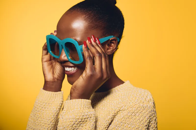 Happy African woman with dark skin wearing large blue sunglasses and yellow sweater smiles against bright yellow background. Concept for fashion, joy, and summer style.
