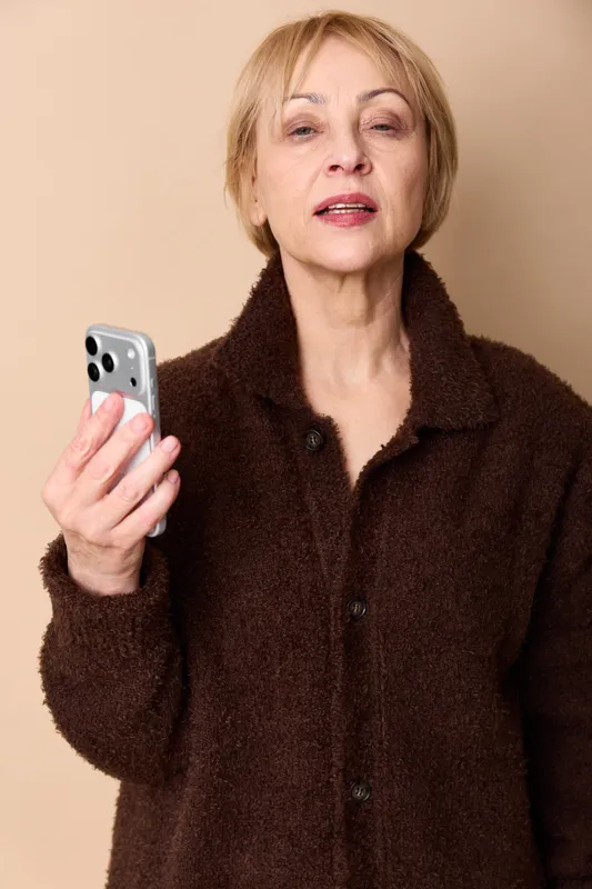 Older woman portrait indoors, holding smartphone, making video call, calm confident expression, wearing brown coat, beige background, close up lifestyle concept, communication