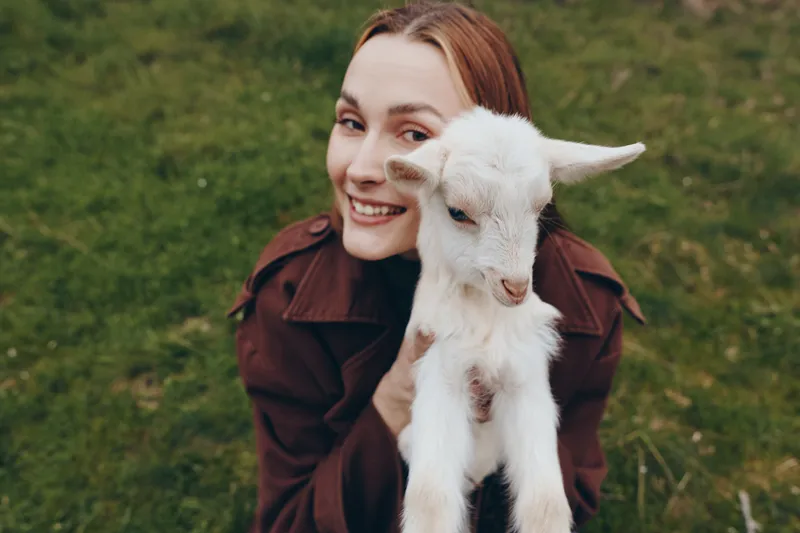 Young woman with red hair holds a white baby goat outdoors smiling warmly in a casual brown jacket on green grass, capturing a joyful lifestyle moment with nature and animals.