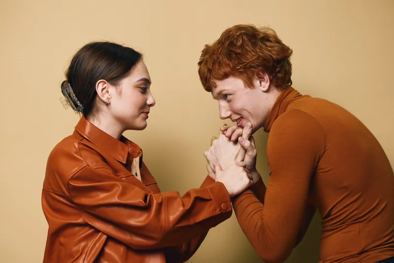 Couple holding hands and looking at each other with affectionate expressions in casual clothing on neutral background. Young man and woman showing trust and connection indoors.