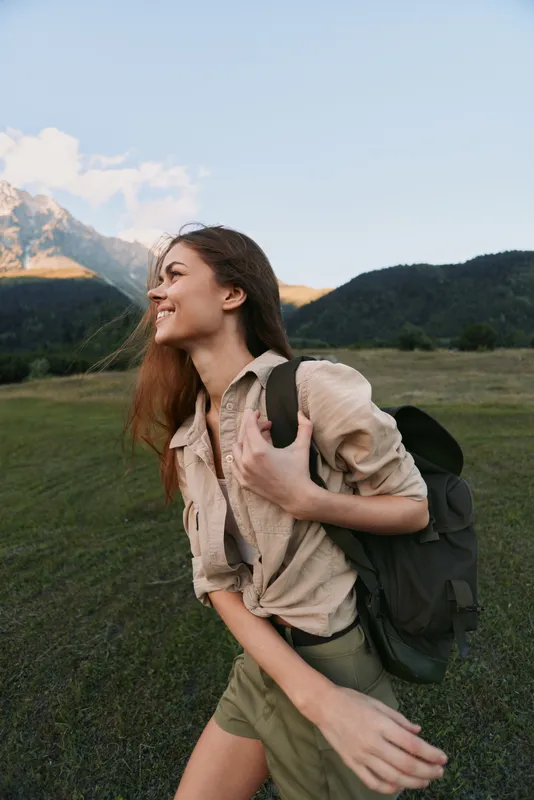 Cheerful woman outdoors, hiking, backpack, sunlit meadow with mountains, casual attire, carefree smile, natural colors, fresh air, travel mood in open valley, breeze, joy, wellbeing, freedom