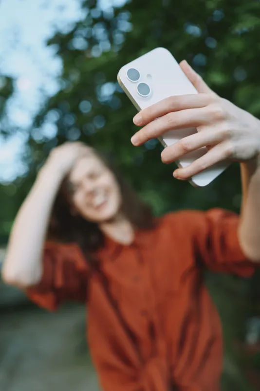 Woman lifestyle portrait outdoors taking a casual selfie with smartphone, relaxed mood, natural light, candid moment, park setting, cheerful expression, everyday style