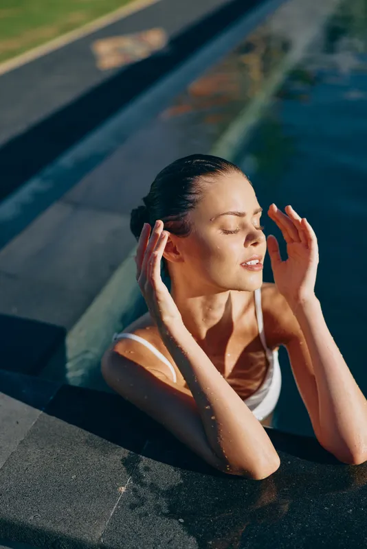 Relaxed woman in a sunlit pool holds a peaceful pose with closed eyes and hands near her face, enjoying warm breeze, casual summer mood, outdoor water setting, serene moment, lifestyle.