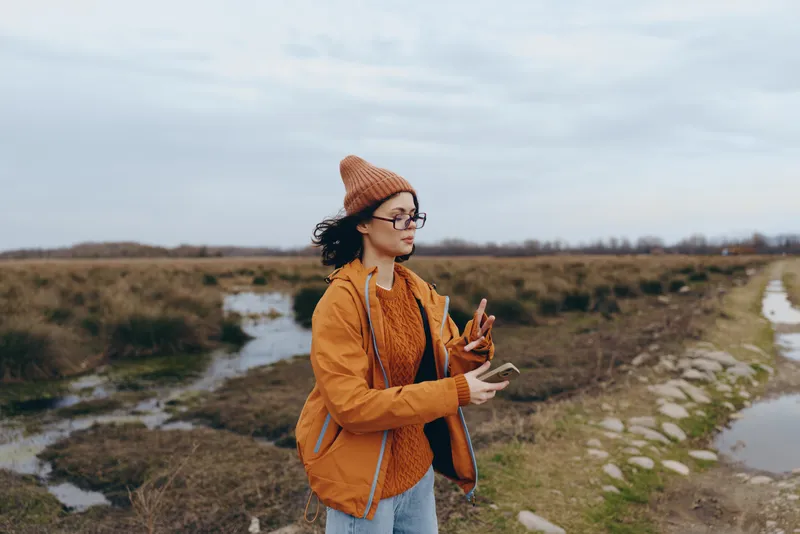 Woman lifestyle portrait outdoors, countryside path under cloudy sky, wearing orange jacket, beanie and glasses, winter feel, gesturing with hands, calm confident mood, scenic nature background