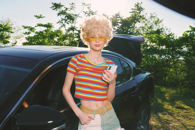 Young woman with retro style stands by a car in a sunny outdoor setting, wearing a striped rainbow top and sunglasses, holding a smartphone, embodying casual lifestyle and vintage film tones.