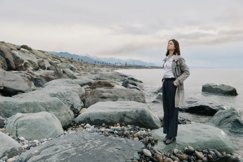 Young woman in casual clothes stands on rocky shore, looking at sea with mountains in background. Cloudy sky creates calm mood. Concept of nature, travel, and contemplation.