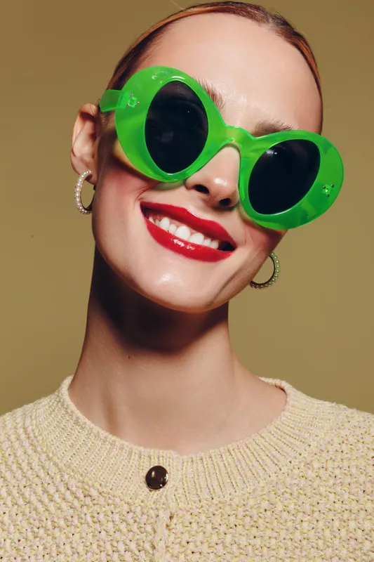 Smiling woman with bright red lipstick wearing large green sunglasses and hoop earrings. She wears a beige knitted sweater against a neutral background. Fashion and style concept.