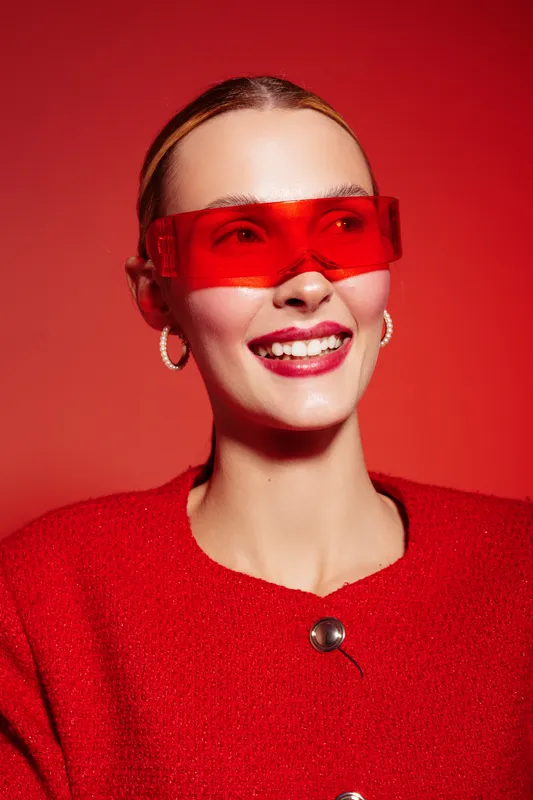 Smiling woman with red sunglasses and red sweater poses on red background. Fashion portrait with bright colors and modern accessories. Concept of style, confidence, and beauty.